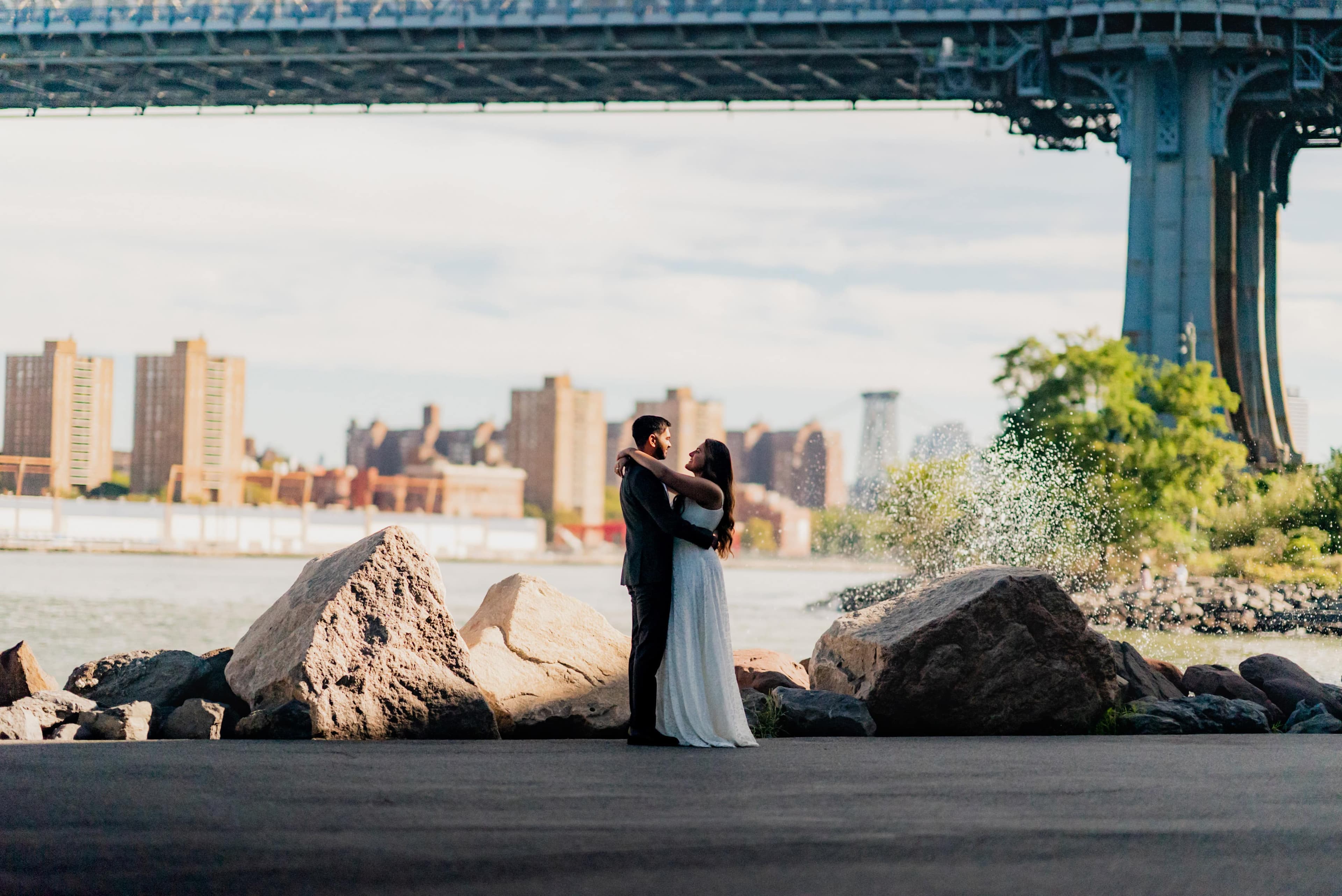 Engaged couple embracing under the Manhattan Bridge at golden hour with the New York City skyline behind them